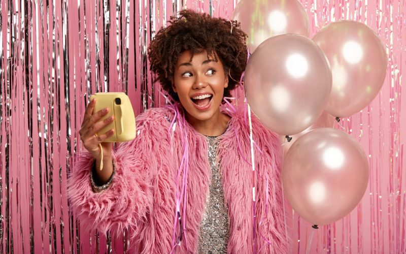 Playful black ethnic woman makes selfie with toy camera, has trendy makeup, prepares for party, holds air balloons, dressed in fur coat and sparkling dress over pink background, has best night ever