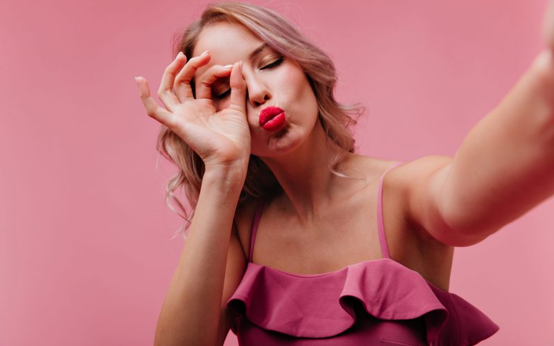 Romantic girl with red lips posing with kissing face expression during indoor photoshoot. Appealing curly woman in pink dress making selfie